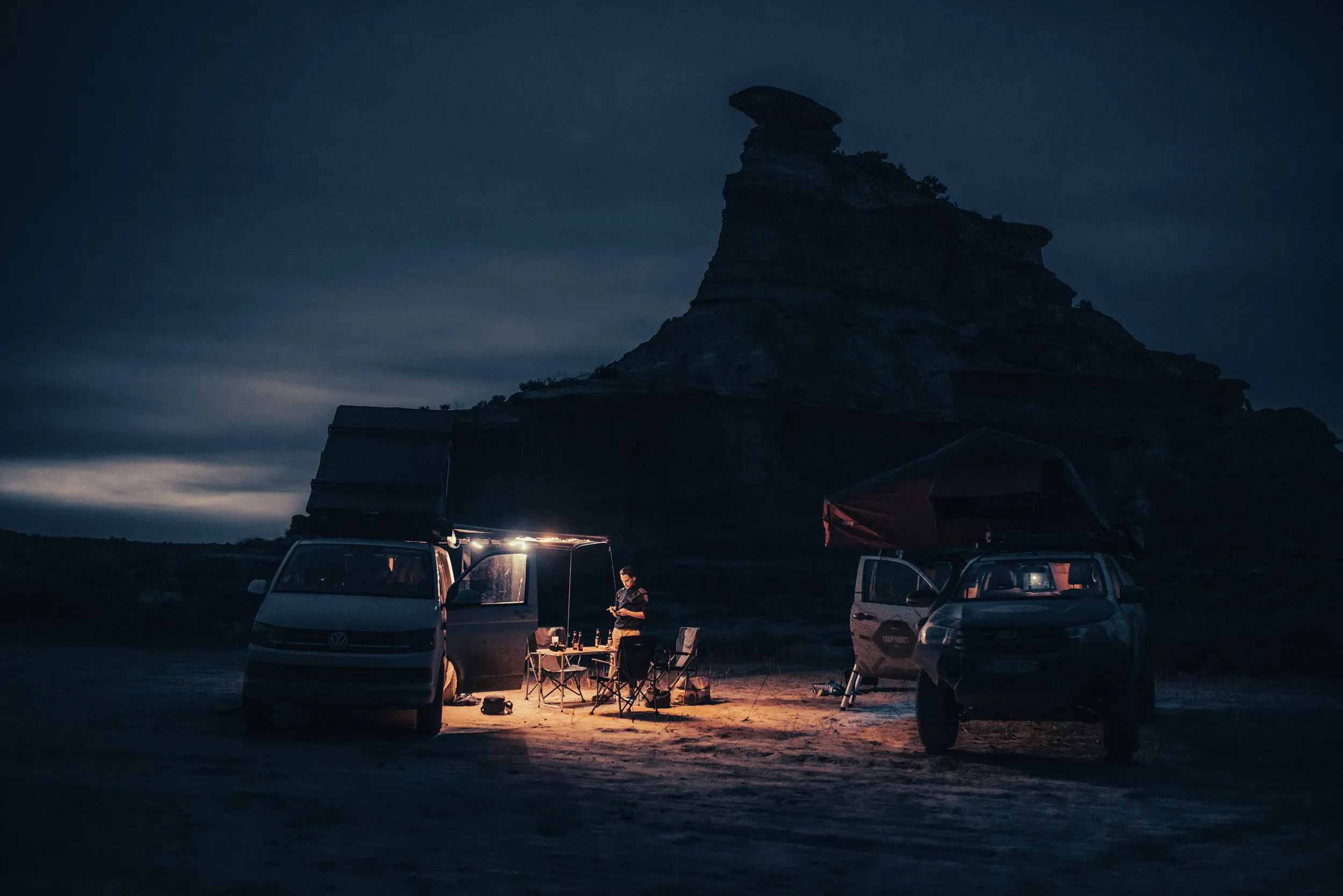 Bivouac de nuit entre un van et un 4x4, éclairé par une lanterne au pied d'une butte rocheuse