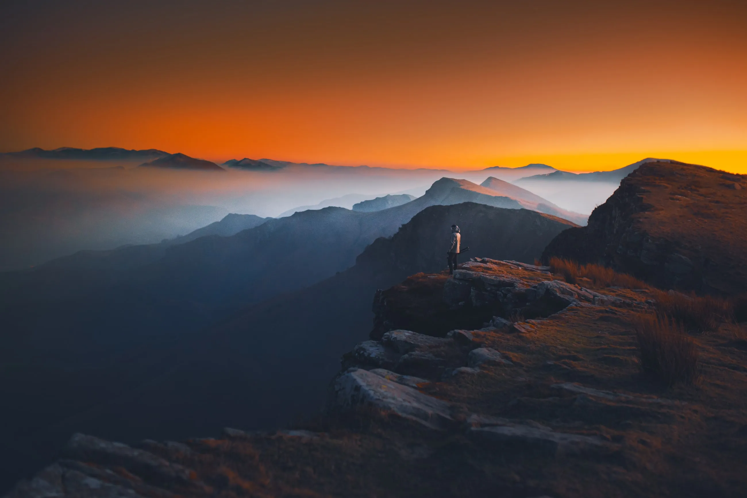 Silhouette contemplant une mer de nuages au coucher du soleil, depuis un éperon rocheux en montagne