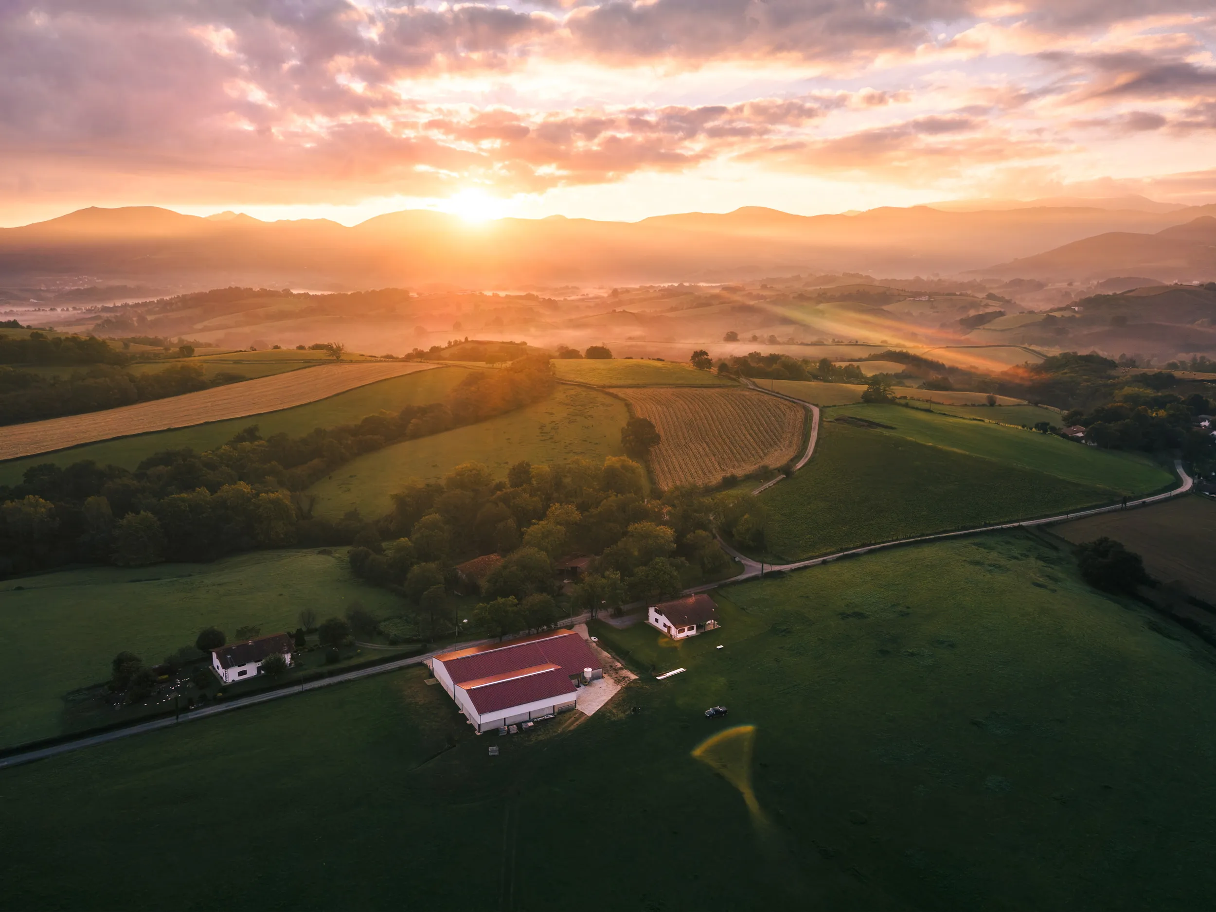 Vue aérienne au drone d'une ferme baignée par le soleil levant, montagnes à l'horizon