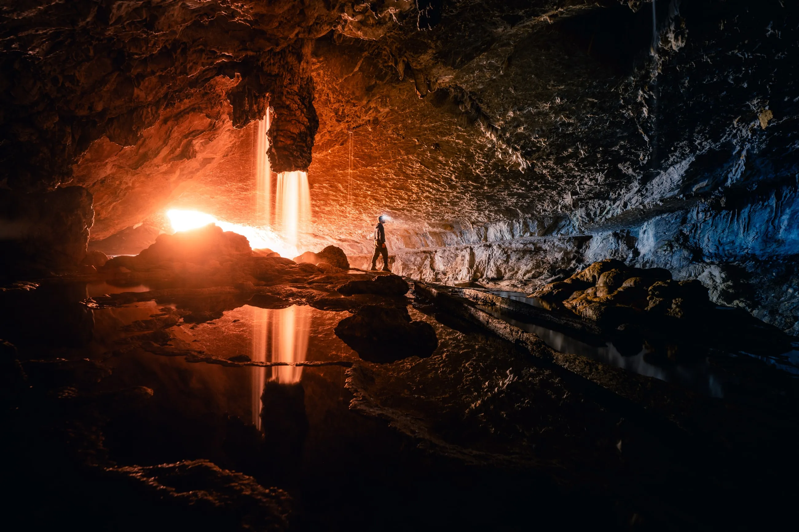 Silhouette explorant une grotte éclairée par une cascade traversée de lumière dorée dans les Pyrénées