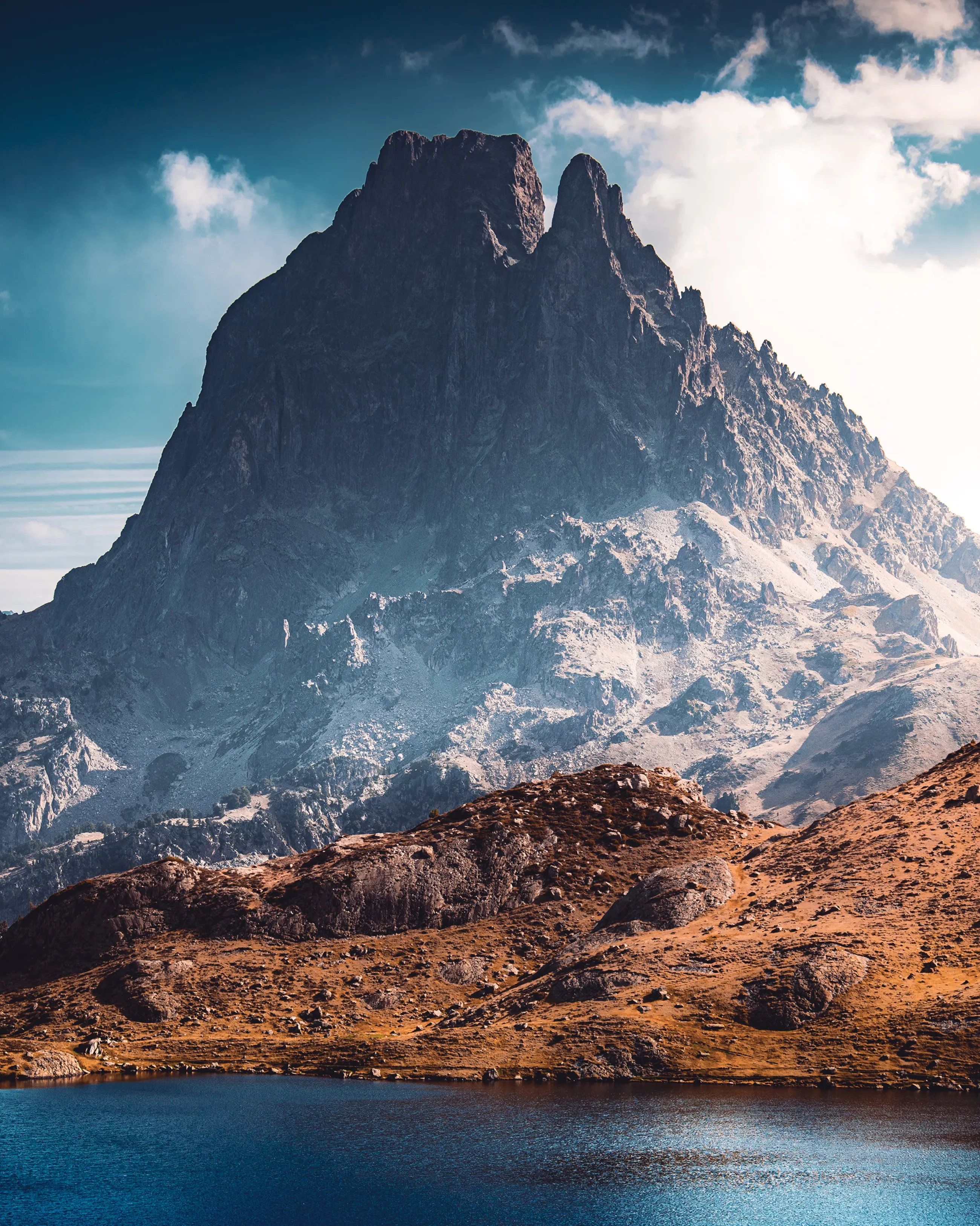 Le pic du Midi d'Ossau sous les nuages blancs, ses pentes enneigées dominant un lac et des rochers fauves
