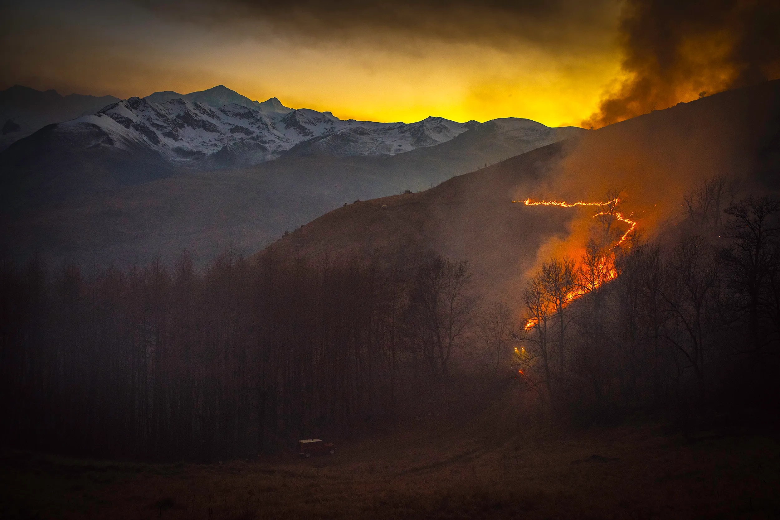 Une ligne de feu d'écobuage serpente sur la montagne au crépuscule, face aux sommets enneigés de Luchon