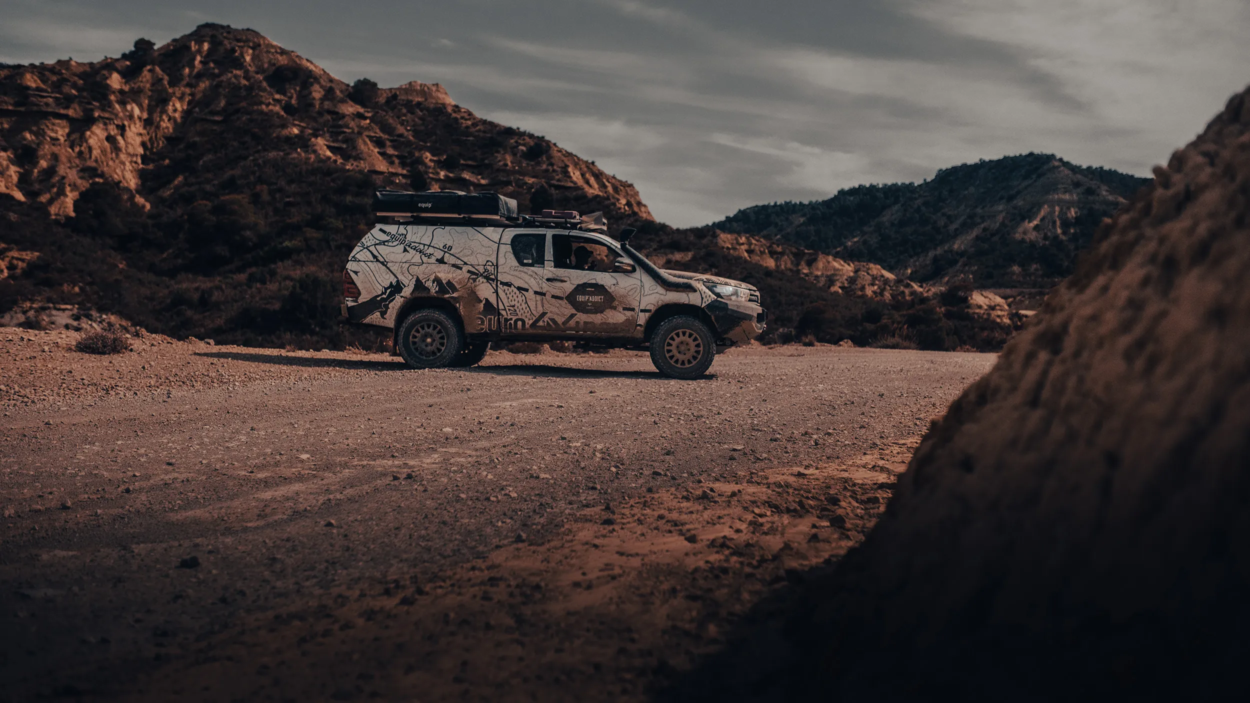 Toyota Hilux avec tente de toit sur une piste désertique, entre des reliefs érodés