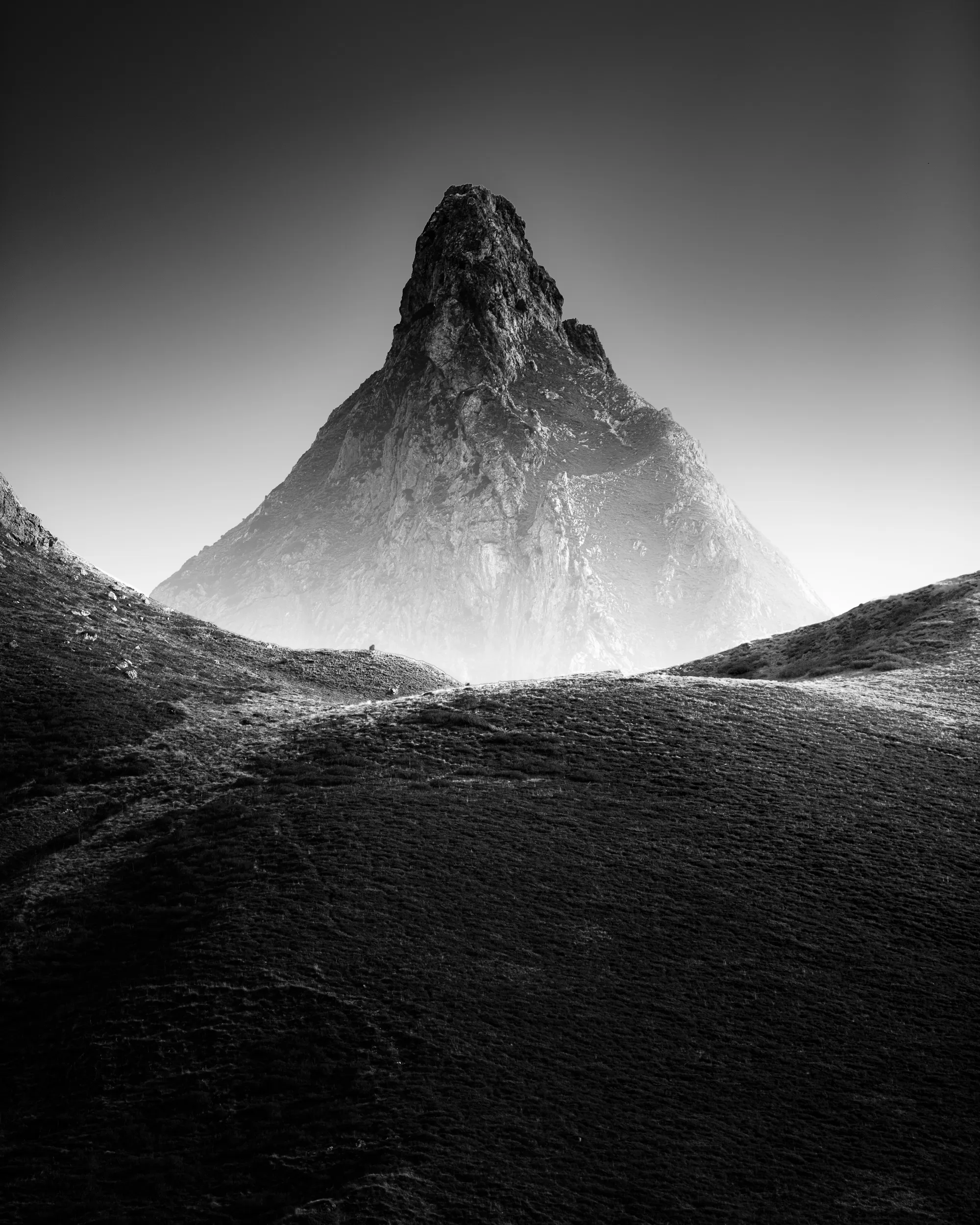 Un sommet pointu émerge de la brume au-dessus de collines ondulantes, en noir et blanc, vallée d'Ossau